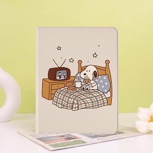 Op de afbeelding: Een beige notitieboek met een cartoonillustratie van een witte hond met bruine oren die in een houten bed zit, een koekje eet en uit een mok drinkt. Naast het bed staat een bruine televisie op een houten nachtkastje met sterren.