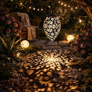 Puede incluir: Una linterna colgante con energía solar y un diseño floral proyecta un patrón de luz sobre un camino de piedra. La linterna cuelga de una rama de árbol en un jardín, con una silla de mimbre y luces de cadena.