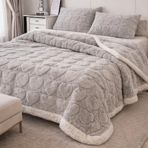 Puede incluir: Una colcha gris de felpa y dos fundas de almohada a juego con un estampado de corazones. La colcha tiene una superficie suave y texturizada y un borde de borreguillo blanco. La ropa de cama está sobre una cama con una alfombra de color neutro.