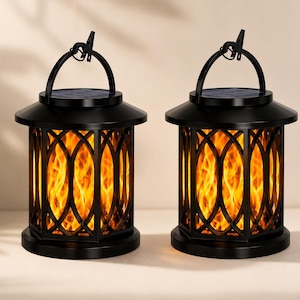 May include: Two black solar-powered lanterns featuring a hexagonal frame with an arched lattice design. Each lantern has a top handle and emits a warm, flickering orange light that mimics the appearance of a real flame inside the glass panels.