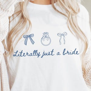 May include: White t-shirt featuring blue line drawings of a bow, a ring, and a bow. The words "Literally just a bride" are written in blue script below the illustrations. This t-shirt is part of a bridal collection.