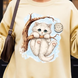 May include: A light yellow sweatshirt featuring a graphic of a white kitten with blue eyes hanging from a tree branch. The kitten is surrounded by a light blue background. The sweatshirt has a round neck and long sleeves.