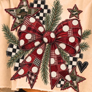 May include: A beige cropped sweatshirt featuring a large central graphic of a red plaid bow with white polka dots, surrounded by green pine needles, black and white checkered squares, camouflage stars, and a small black heart outline.