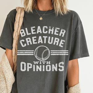 Puede incluir: Camiseta gris oscuro con el texto "BLEACHER CREATURE WITH OPINIONS" en letras blancas desgastadas. Un gráfico de béisbol está centrado entre las palabras. La camiseta es de estilo casual, adecuada para el uso diario.