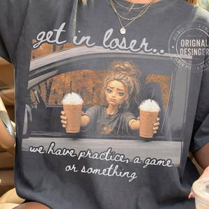 Op de afbeelding: Een antracietgrijs t-shirt met een afbeelding van een persoon in een auto die twee ijskoffies vasthoudt. De tekst luidt, get in loser... we have practice, a game of something. Het ontwerp bevat een cirkelvormig logo in de rechterbovenhoek.