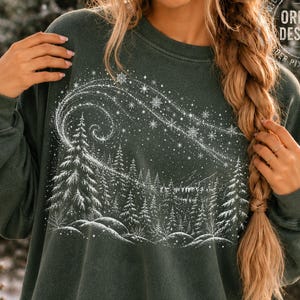 May include: A dark green long-sleeve sweatshirt featuring a white graphic print of a winter forest scene with snow-covered pine trees and a swirling starry sky with snowflakes above the treeline, worn with blue denim jeans.