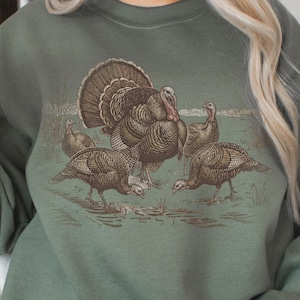 May include: A sage green sweatshirt featuring a vintage-style illustration of a male turkey with its tail feathers fanned out, surrounded by several smaller turkeys. The design is in shades of brown and tan.