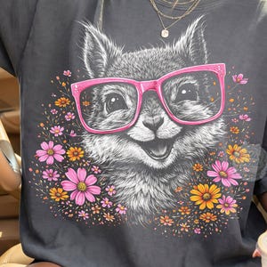 May include: Dark gray t-shirt featuring a smiling fox wearing pink glasses, surrounded by pink, orange, and yellow flowers. The graphic tee has a relaxed fit and is made of soft material.
