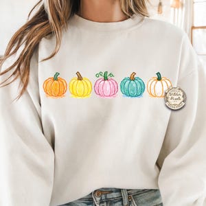 May include: Cream-colored sweatshirt featuring a row of six colorful pumpkins in orange, yellow, pink, teal, and white. The pumpkins are outlined in black and have green stems. The sweatshirt has a round neck and long sleeves. The text "Wilder Pixels" is on the right side.