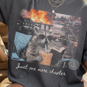 Peut inclure: T-shirt gris foncé avec un dessin d'un raton laveur portant des lunettes de soleil, assis dans une chaise de plage, buvant et lisant. L'image comprend un bâtiment en feu et le texte "Just one more chapter".