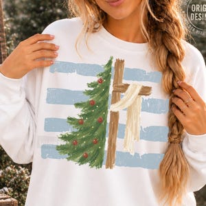 May include: A white long-sleeve sweatshirt featuring a graphic print of a green Christmas tree on the left and a wooden cross draped with a white cloth on the right, set against a background of horizontal light blue distressed stripes.