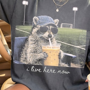 May include: Dark gray t-shirt featuring a raccoon wearing sunglasses and a denim cap, drinking from a clear cup with a straw. The raccoon is sitting in a chair in front of a soccer field. The text "i live here now" is printed below the image.