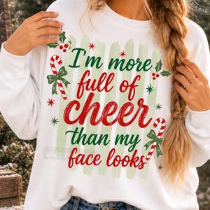 May include: A white long-sleeve sweatshirt featuring a green brushstroke background and red and green text that reads, I'm more full of cheer than my face looks. The design includes red and white candy canes, green bows, and small red berries.