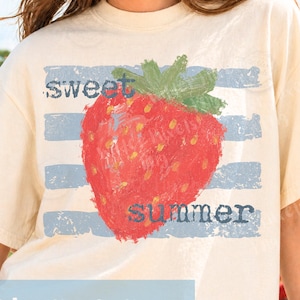 Könnte beinhalten: Cremefarbenes T-Shirt mit einer großen roten Erdbeer-Grafik und den Wörtern "sweet" und "summer" in Blau. Das Design umfasst blaue horizontale Streifen. Ein lässiges Sommertop.