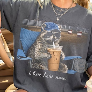 May include: Dark gray t-shirt featuring a raccoon wearing sunglasses and a denim cap, seated in a blue folding chair, sipping an iced coffee. The text "i live here now" is printed below the raccoon.