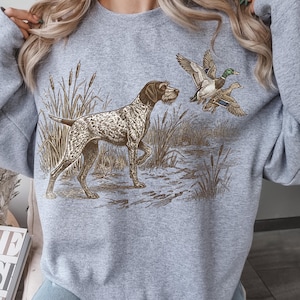 Peut inclure: Un sweat-shirt gris clair avec une scène de chasse. Le motif représente un chien marron et blanc, des roseaux et deux canards en vol. Le sweat-shirt a des manches longues et un col rond.