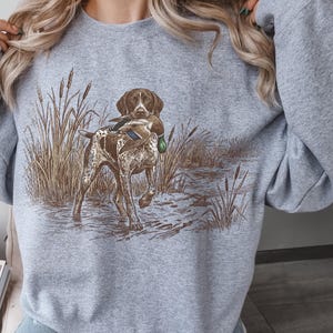 May include: A light gray sweatshirt featuring a hunting dog carrying a duck in its mouth. The dog is brown and white, standing in water with tall grass in the background. The design is a detailed illustration.