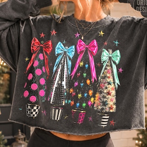 May include: A charcoal gray long-sleeve cropped sweatshirt featuring a graphic print of four stylized Christmas trees topped with large bows in red, blue, pink, and teal. The trees are decorated with colorful lights, stars, and patterns.