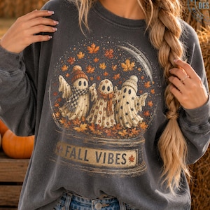Könnte beinhalten: Ein anthrazitfarbenes Sweatshirt mit Rundhalsausschnitt und einer Grafik von drei Geistern in einer Schneekugel, die mit herbstlichen Blättern gefüllt ist. Die Geister tragen Strickmützen und Schals. Unter der Kugel steht der Text FALL VIBES.