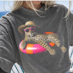 Op de afbeelding: Donkergrijs T-shirt met een luipaard met een zonnebril en een strohoed, liggend op een roze en oranje opblaasbare ring. Het luipaard heeft een gevlekte vacht. Het shirt heeft een relaxte pasvorm.