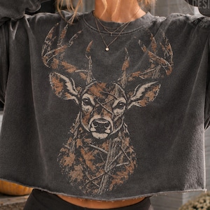 Peut inclure: Un sweat-shirt court gris anthracite orné d'un grand motif détaillé représentant une tête de cerf avec des bois. L'illustration du cerf intègre des motifs de camouflage marron et beige. Le sweat-shirt a des manches longues et un ourlet brut.