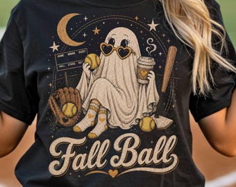 May include: A black t-shirt featuring a graphic of a ghost wearing heart-shaped sunglasses, holding a softball and a coffee cup. The design includes a baseball bat, a scoreboard, a crescent moon, stars, and the text Fall Ball in a script font.