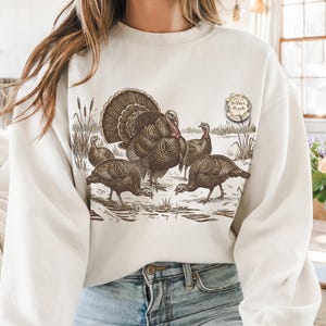 May include: Cream-colored sweatshirt featuring a detailed illustration of a group of turkeys in a winter scene. The design includes a large male turkey with a fanned tail and several smaller turkeys. The text "Wilder Pixels" is on the upper right.