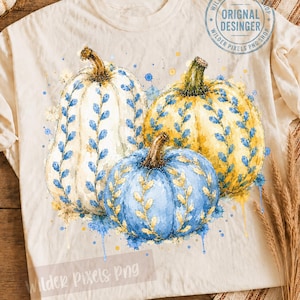 May include: A cream-colored t-shirt featuring a watercolor graphic of three pumpkins. The pumpkins are white, yellow, and blue, decorated with blue and gold leaf patterns. The text "ORIGINAL DESINGER" and "WILDER PIXELS PNG" is visible.