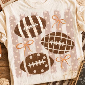 Könnte beinhalten: Ein cremefarbenes T-Shirt mit einem Grafikdruck von drei braunen Footballs mit weißen Schnürsenkeln. Die Footballs sind mit Streifen, einem Gittermuster und Punkten verziert, durchsetzt mit drei orangefarbenen, liniengezeichneten Schleifen auf einem Hintergrund aus vertikalen hellbraunen Pinselstrichen und weißen Punkten.