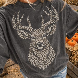 May include: A charcoal gray short-sleeve t-shirt featuring a large graphic print of a deer head. The deer is rendered in a beige and black leopard print pattern, covering the entire head and antlers, set against the solid dark fabric of the shirt.