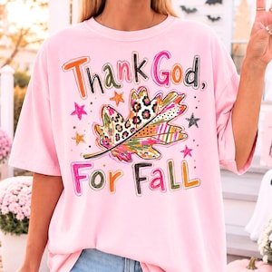 Puede incluir: Una camiseta de manga corta de color rosa claro con el texto Thank God, For Fall en fuentes coloridas y variadas. El gráfico central muestra una hoja de otoño grande con una mezcla de estampado de leopardo, rayas y flores, rodeada de estrellas.