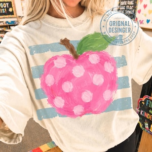 Op de afbeelding: Een beige T-shirt met korte mouwen voorzien van een grote, roze, gestippelde appelafbeelding met een groen blaadje en een bruine steel. De appel is gecentreerd over horizontale, verweerde lichtblauwe strepen. Een cirkelvormig watermerklogo is rechtsboven zichtbaar.