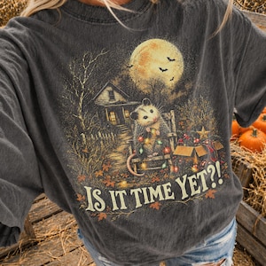 Könnte beinhalten: Ein anthrazitfarbenes T-Shirt mit einem Grafikdruck eines Opossums, das auf einem mit Lichterketten geschmückten Stuhl vor einem Haus unter einem Vollmond sitzt. Der Text "IS IT TIME YET?!" ist unten in einer Schriftart aufgedruckt.