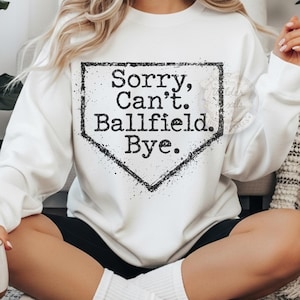 May include: White crew neck sweatshirt featuring the text "Sorry, Can't. Ballfield. Bye." in a black, distressed font within a home plate graphic. The sweatshirt is a casual style.