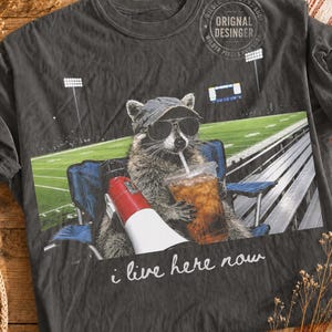 Może przedstawiać: Ciemnoszary t-shirt z grafiką przedstawiającą szopa pracza w czapce z daszkiem i okularach przeciwsłonecznych, siedzącego na niebieskim składanym krześle na trybunach. Szop trzyma megafon i kubek. Napis na koszulce brzmi: i live here now.