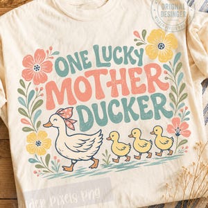 May include: A cream-colored t-shirt featuring a graphic print of a mother duck with a bow and three ducklings. The text reads ONE LUCKY MOTHER DUCKER in teal and coral. The design is framed by colorful flowers and green leaves in a retro style.