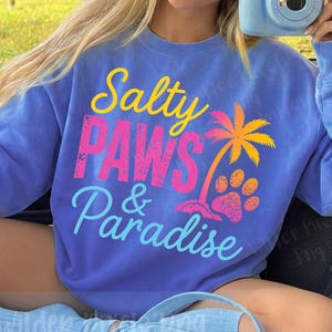 Könnte beinhalten: Ein blaues Sweatshirt mit den Worten "Salty Paws & Paradise" in Gelb, Pink und Blau. Auf der Vorderseite des Shirts befinden sich außerdem eine Palmen- und Pfotenabdruck-Grafik. Das Sweatshirt ist ein bequemer, lässiger Stil.