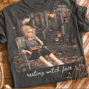 May include: A charcoal grey t-shirt featuring a graphic of a person wearing a witch hat, sitting in a lawn chair holding a bowl of sweets. The background shows a house with pumpkins and the text "resting witch face" in white script.