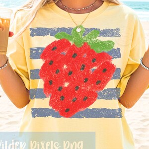Op de afbeelding: Geel T-shirt met een grote rode aardbei-afbeelding en blauwe horizontale strepen. De aardbei heeft zwarte zaden en groene bladeren. De tekst "Wilder Pixels Png" staat onderaan.
