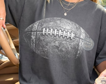 May include: Dark grey t-shirt featuring a distressed, grayscale football graphic. The image of the football is centered on the shirt. The shirt has a relaxed fit and short sleeves. The shirt is a casual style.