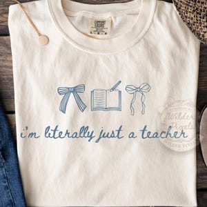 Op de afbeelding: Crèmekleurig T-shirt met blauwe illustraties van strikken, een open boek en een pen. De tekst "I'm literally just a teacher" is in blauwe letters gedrukt. Het shirt heeft een ronde hals en een Comfort Colors label.