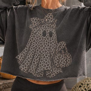 Peut inclure: Un sweat-shirt court gris anthracite avec un motif de fantôme portant un nœud et tenant une citrouille. Le fantôme et la citrouille présentent un imprimé léopard noir et gris. Le sweat-shirt a un col rond et des bords bruts à l'ourlet.