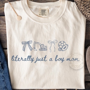 May include: Cream-colored t-shirt with the phrase "literally just a boy mom" in blue script. The shirt features blue line drawings of a bow, excavator, bow, soccer ball, and football. The shirt is a Comfort Colors brand.