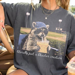 May include: Dark gray t-shirt featuring a raccoon wearing sunglasses and a denim cap, holding a drink with a straw. The graphic includes the text "literally just a bleacher creature". The raccoon is sitting in a chair with a baseball field in the background.