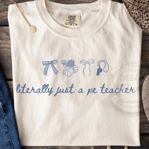 May include: Cream-colored t-shirt with blue illustrations of sports equipment and the phrase "literally just a pe teacher." The shirt features a crew neck and short sleeves. The shirt is made of a soft, comfortable material.