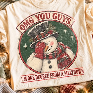 May include: A cream-coloured t-shirt featuring a graphic of a snowman wearing a black top hat and red plaid scarf, covering its face with a red mitten. The text reads "OMG YOU GUYS, I'M ONE DEGREE FROM A MELTDOWN" in red letters within a circle.