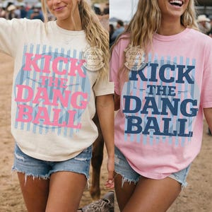 May include: Two t-shirts, one cream and one pink, each with the text "KICK THE DANG BALL" in pink and blue. The shirts are worn with denim shorts. The shirts have a round graphic design with vertical stripes.