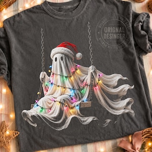 Könnte beinhalten: Ein anthrazitfarbenes T-Shirt mit einer Grafik eines weißen Geistes, der eine rot-weiße Weihnachtsmütze trägt und auf einer Holzschaukel sitzt. Der Geist ist mit bunten Lichterketten umwickelt. Der Text "ORIGINAL DESINGER" ist oben rechts aufgedruckt.
