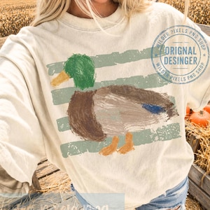 Pu&ograve; includere: T-shirt color crema con un disegno di anatra dipinta con accenti verdi, marroni e blu. L'anatra &egrave; posizionata su strisce orizzontali verde chiaro. La maglietta ha il testo "ORIGINAL DESIGNER" e "WILDER PIXELS PNG SHOP" in un logo circolare.