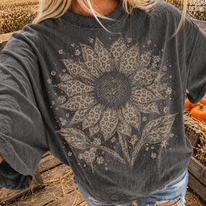 May include: A charcoal gray short-sleeve t-shirt featuring a large, central sunflower graphic. The sunflower petals are filled with a tan leopard print pattern. The design includes small scattered dots and splatters around the flower.